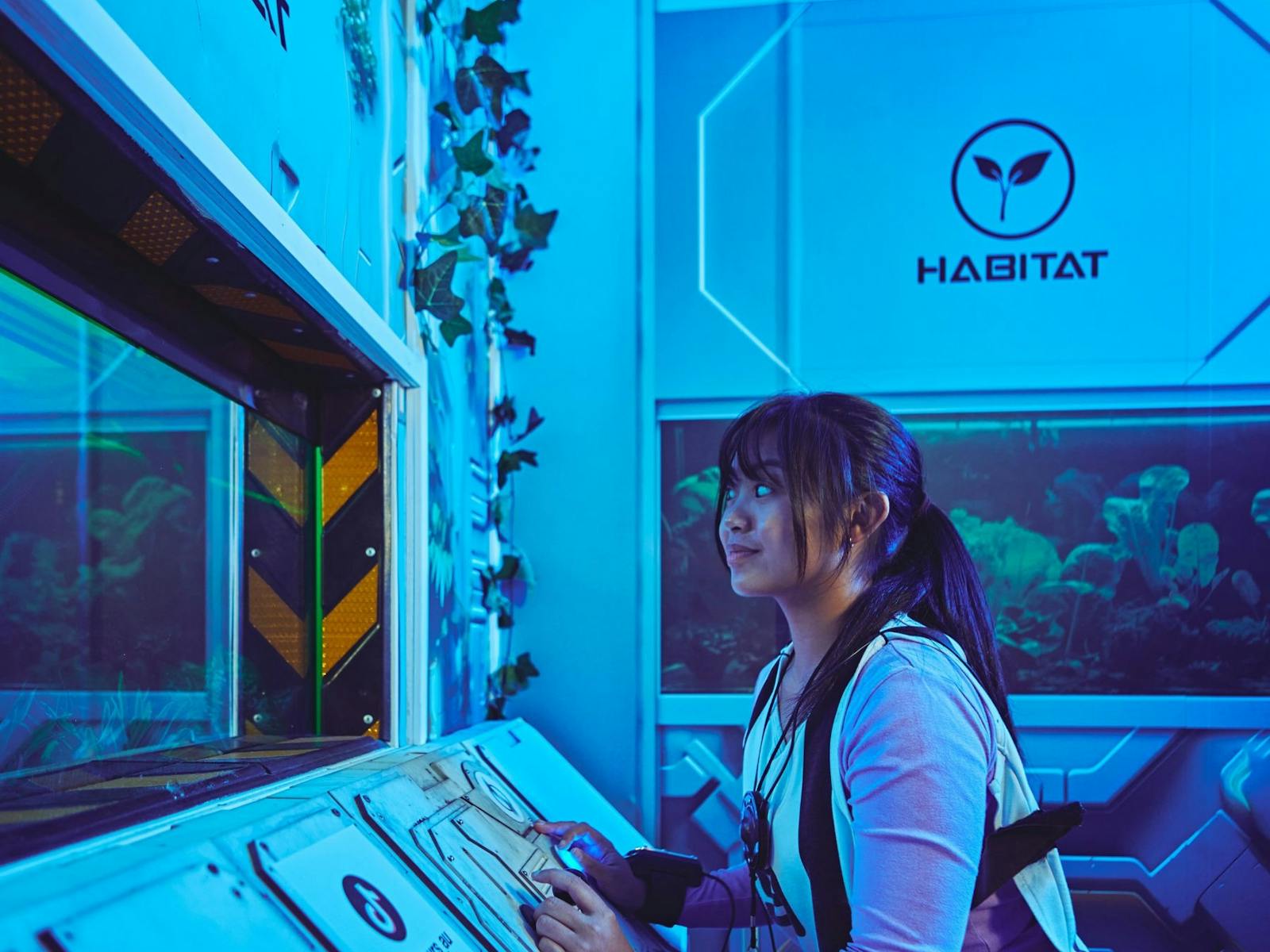 A young woman in a Saboteurs vest works at a control panel in a blue-lit Habitat room.